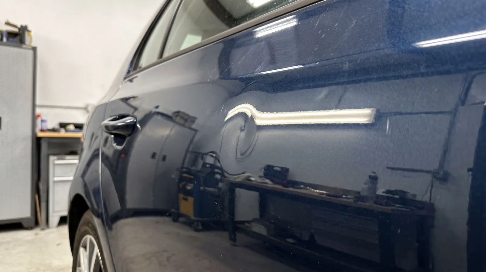 A round dent in the rear door of a dark blue car, a strip light bent round it in the paint