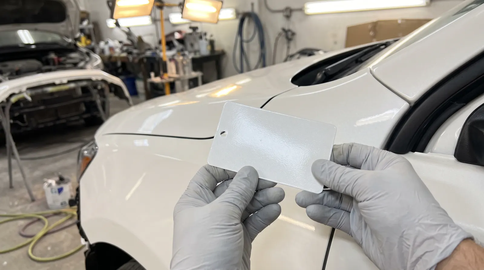 Gloved hands holding a sprayed white test card against the wing of a white car to check the colour match