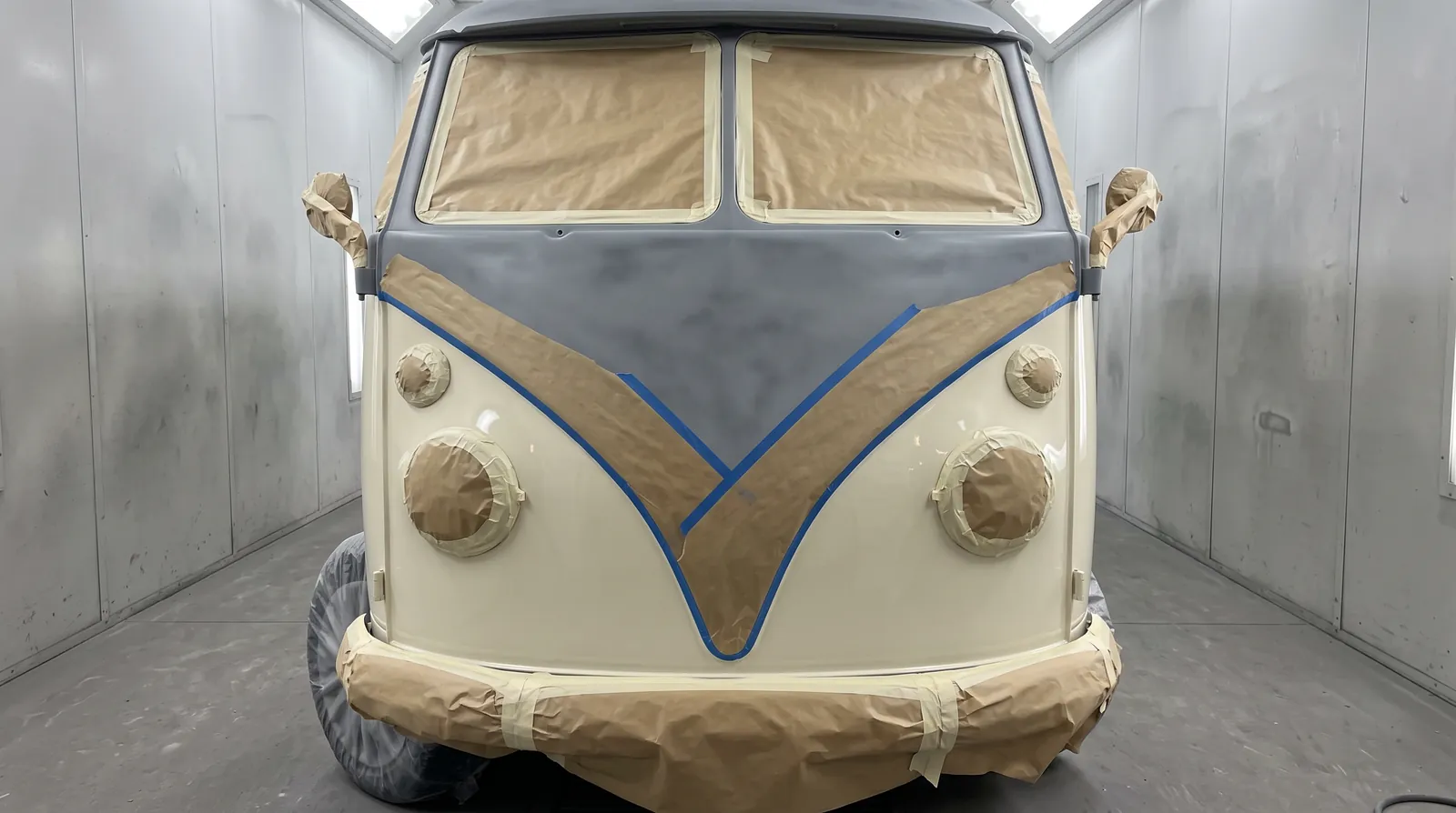 A split-screen camper's front in the spray booth, the cream done and the V of the split line laid out in blue tape and masking paper, the top still in grey primer