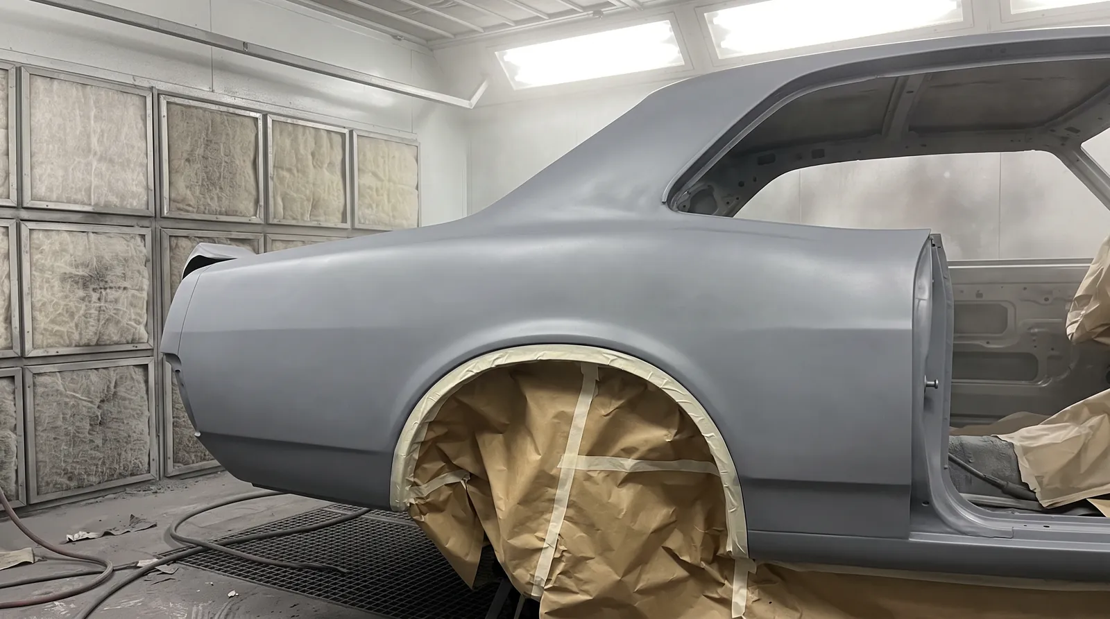 A classic coupe shell in grey primer in the spray booth, its wheel arches masked off