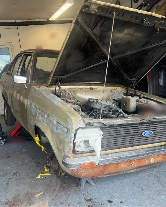 A gold Mk2 Escort in the shop with its bonnet up, rust along the front valance and bare patches on the wing