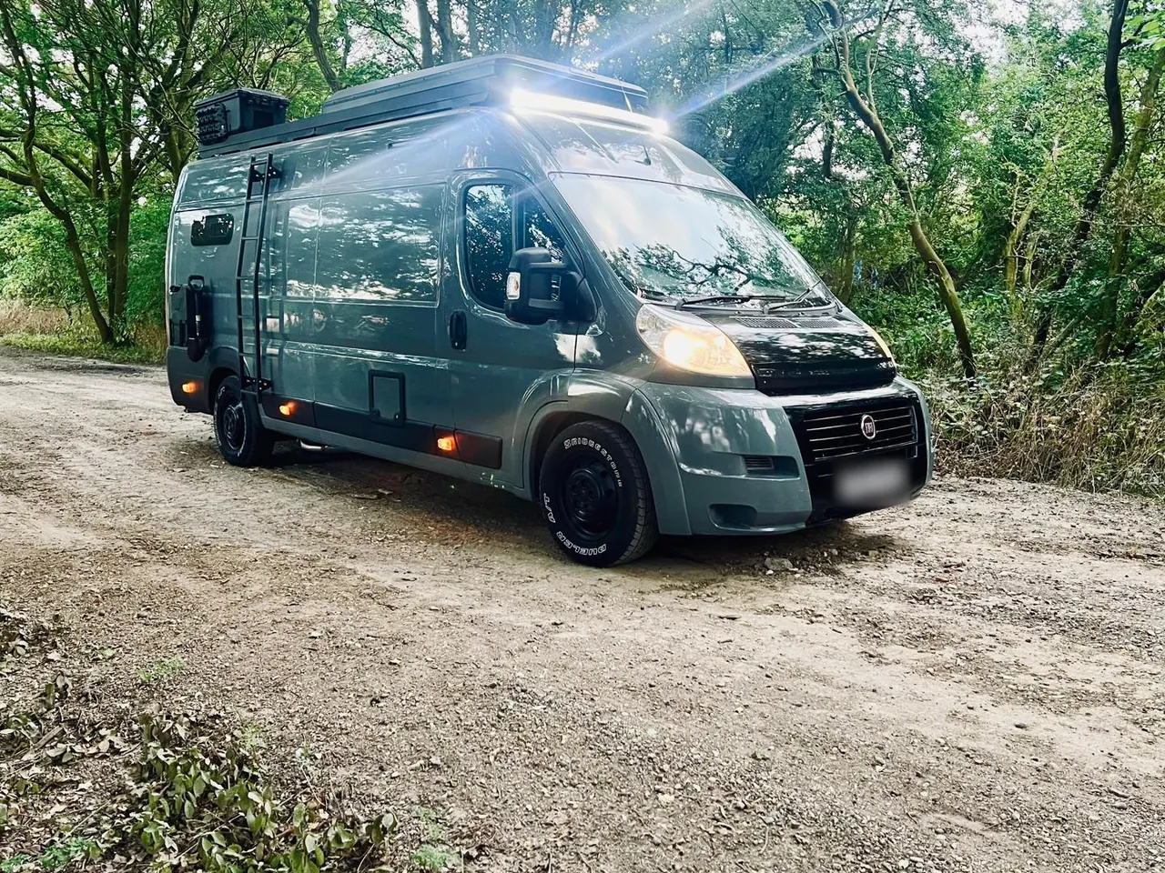A grey Fiat Ducato camper van with a roof rack and light bar on a woodland track